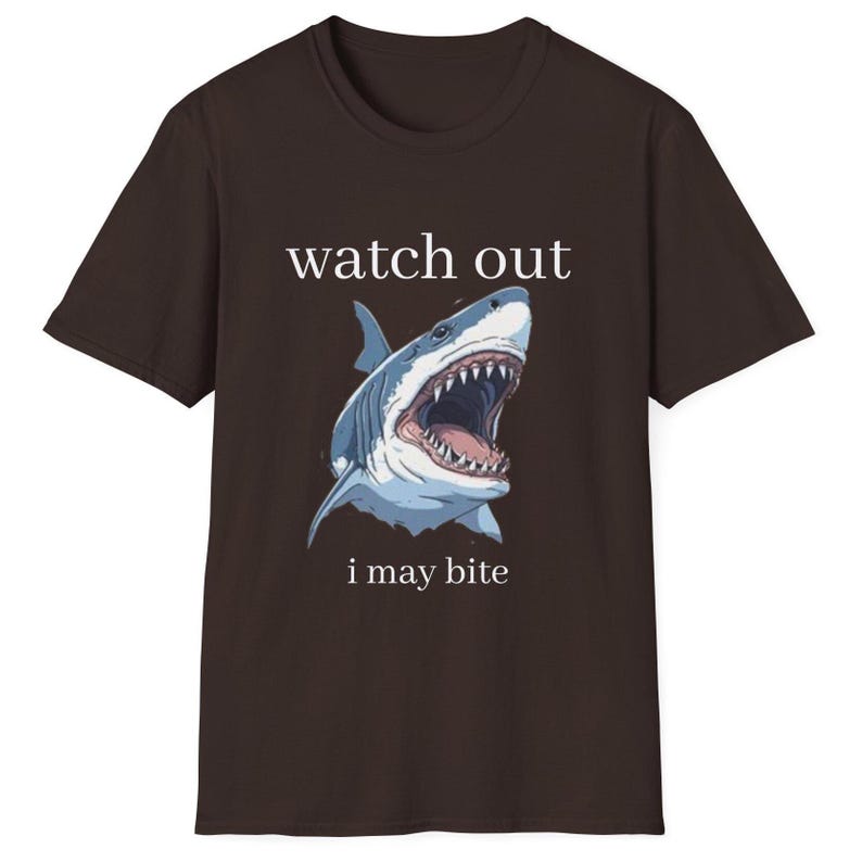May include: A brown t-shirt featuring a detailed illustration of a shark with an open mouth, displaying sharp teeth. The text "watch out i may bite" is printed above and below the shark image.