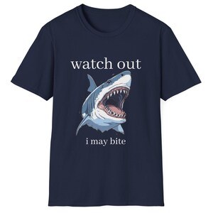 May include: Navy blue t-shirt featuring a detailed illustration of a shark with an open mouth, teeth visible. The text "watch out i may bite" is printed above and below the shark graphic. The shirt is a casual wear item.