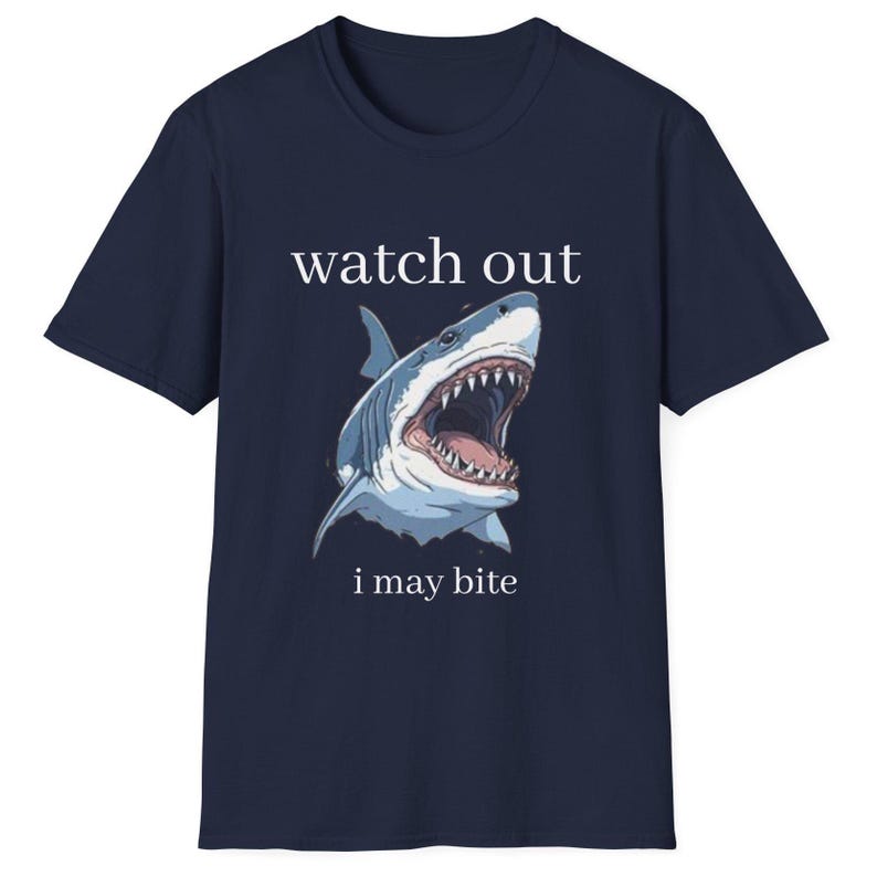 May include: Navy blue t-shirt featuring a detailed illustration of a shark with an open mouth, teeth visible. The text "watch out i may bite" is printed above and below the shark graphic. The shirt is a casual wear item.