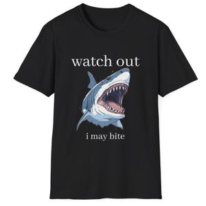 May include: Black t-shirt featuring a detailed illustration of a shark with an open mouth, showcasing sharp teeth. The text "watch out" is above the shark, and "i may bite" is below. The shark is colored in shades of blue and white.