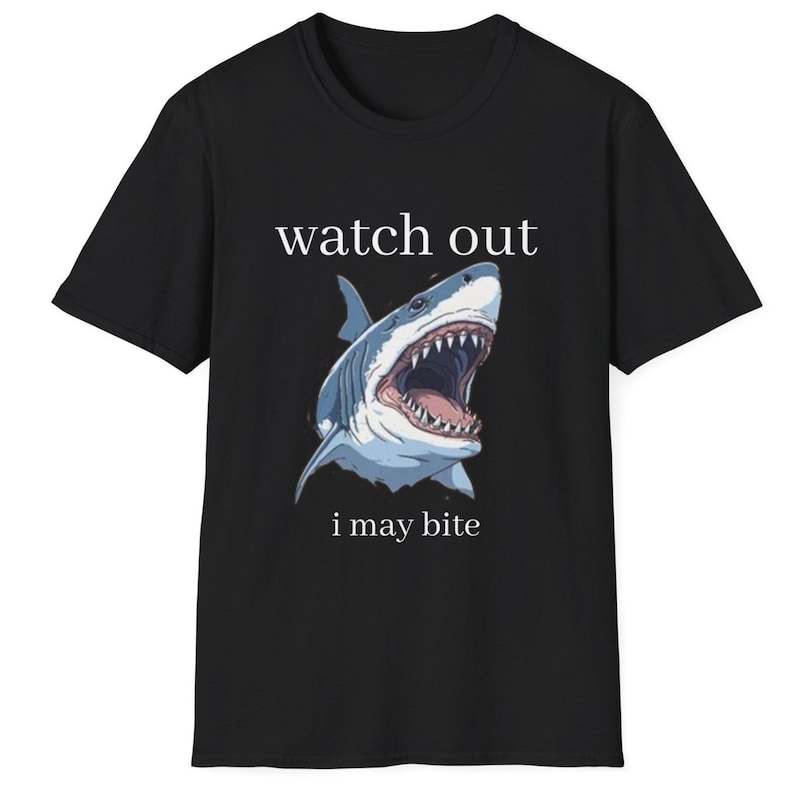 May include: Black t-shirt featuring a detailed illustration of a shark with an open mouth, showcasing sharp teeth. The text "watch out" is above the shark, and "i may bite" is below. The shark is colored in shades of blue and white.