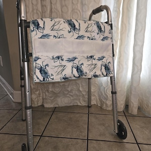 Op de afbeelding: Een zilveren rollator met een blauw-wit patroon. De tas heeft een herhalend ontwerp van krabben en bladeren. De rollator heeft zwarte handvatten en wielen, voor een wit gordijn.