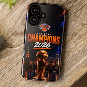 May include: A black smartphone case featuring a New York Knicks logo, the text NEW YORK CHAMPIONS 2026, and a graphic of a golden championship trophy set against a dark city skyline at night with orange and blue light accents.