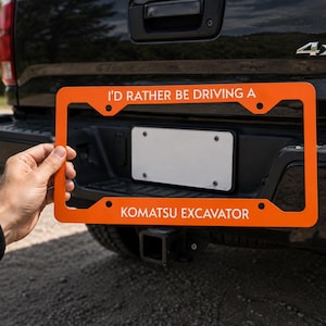 May include: A bright orange license plate frame featuring white text that reads I'D RATHER BE DRIVING A KOMATSU EXCAVATOR. The rectangular frame has four mounting holes and is designed to fit around a standard vehicle license plate.
