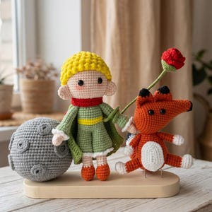 May include: A handmade crochet set featuring a small figure with yellow hair, a green outfit, and a red scarf, standing next to a gray textured sphere and an orange fox holding a red flower, all mounted on a smooth, light-colored wooden base.