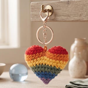 May include: A heart-shaped keyring featuring a multi-coloured crochet design with horizontal stripes in red, orange, yellow, green, blue, and purple. The heart is attached to a metallic rose gold-toned lobster clasp and a circular split ring.