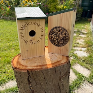 Könnte beinhalten: Zwei helle Holznistkästen mit dunkelgrünen Schrägdächern. Der vordere Nistkasten hat ein kreisförmiges Einflugloch und den eingravierten Text "Welcome to my new home". Der seitliche Nistkasten zeigt eine kreisförmige Gravur zweier Vögel.