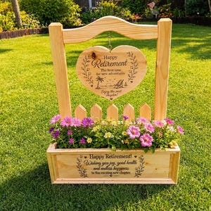 May include: A light-coloured wooden planter box with a picket fence back and a hanging heart-shaped sign. The sign reads Happy Retirement, The best time for new adventures, Enjoy the next chapter. The planter box features the text Happy Retirement! Wishing you joy, good health and endless happiness in this exciting new chapter. The planter is filled with pink and yellow flowers.