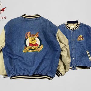 Könnte beinhalten: Zwei Collegejacken aus Jeansstoff. Eine Jacke hat ein großes, gesticktes Winnie-Puuh-Design auf dem Rücken. Die andere Jacke hat ein kleineres Winnie-Puuh-Design auf der Brust. Beide Jacken haben beige Ärmel und gestreifte Verzierungen.