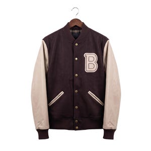 May include: A varsity jacket featuring a dark brown wool-blend body and cream-coloured sleeves. The front includes a large cream-coloured letter B patch on the left chest, a snap-button closure, two side pockets with cream trim, and ribbed cuffs.