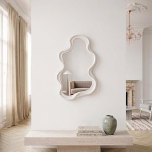 Op de afbeelding: Een wandspiegel met een onregelmatig, golvend wit frame. Onder de spiegel staat een rechthoekige console van beige steen met twee gestapelde boeken getiteld Art & Forme en Design Essentiel, naast een gladde, saliegroene keramische vaas.