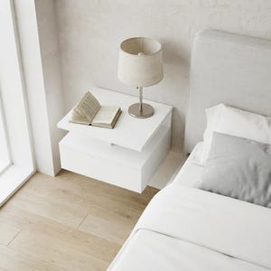 Puede incluir: Una mesita de noche flotante blanca con un cajón y un estante abierto. Una lámpara de mesa de tela beige con base plateada se sitúa sobre la superficie junto a un libro abierto de páginas blancas. La mesita está junto a una cama con ropa blanca.