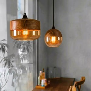 May include: Two pendant lights featuring cylindrical and spherical wooden tops paired with amber-tinted glass shades. Each fixture hangs from a black cord, emitting a warm glow from the internal light bulbs, showcasing natural wood grain textures.