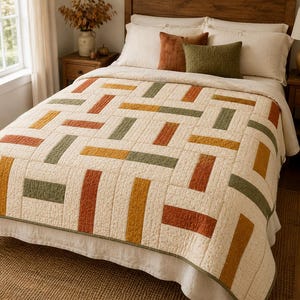 Könnte beinhalten: Eine Steppdecke mit einem geometrischen Muster aus rechteckigen Blöcken in den Farben gebranntes Orange, Senfgelb und Olivgrün auf cremefarbenem Grund mit einer dezent gesprenkelten Textur und sichtbaren linearen Steppnähten.