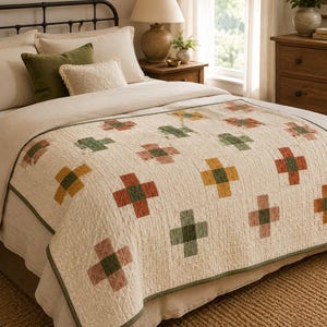 May include: A cream-colored quilt featuring a repeating pattern of cross-shaped motifs in muted shades of terracotta, olive green, and mustard yellow. The quilt has a textured, quilted surface and is finished with a solid olive green fabric border.