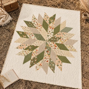 May include: A rectangular quilt featuring a central starburst pattern composed of diamond-shaped fabric patches in solid olive green, solid beige, and a floral print with small orange, pink, and green flowers on a cream background.