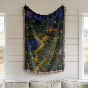 May include: A woven throw blanket featuring a detailed illustration of a cottage at night. The scene includes a path, garden, pond, and starry sky, with warm lighting. The blanket has a fringed edge and is displayed on a white wall.