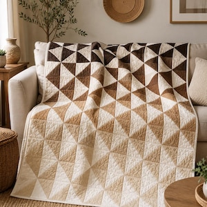 May include: A quilted throw blanket featuring a geometric pattern of triangles in a gradient color scheme. The design transitions from dark brown and tan at the top to light cream and beige at the bottom, with visible stitching throughout the fabric.