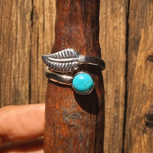 May include: A silver ring featuring a leaf motif and a turquoise gemstone. The ring is presented on a wooden stand, highlighting its delicate design and the vivid colour of the stone. The leaf design adds a touch of nature to the jewellery.