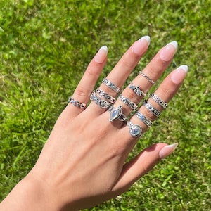 May include: A collection of ten silver-toned rings worn on fingers, featuring various designs including a chain link band, a star-patterned band, a bee motif, a marquise-shaped iridescent stone, and a round iridescent stone set in a decorative frame.