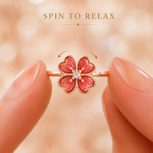 May include: A rose gold-toned ring featuring a four-petaled flower design with red enamel inlay and a central clear stone. The text SPIN TO RELAX is printed above the ring, and curved arrows indicate the flower component is designed to spin.
