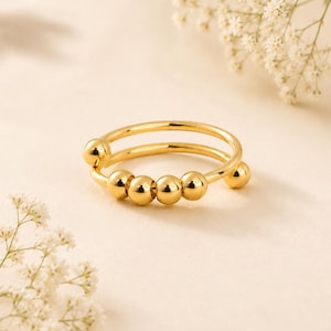 Peut inclure: Une bague dorée dotée d'un design à double anneau avec cinq petites perles sphériques lisses qui coulissent le long du fil. La bague présente une finition polie et réfléchissante ainsi qu'une forme circulaire, soulignant une structure métallique simple.