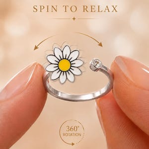Op de afbeelding: Een zilverkleurige metalen ring met een witte en gele madeliefje bedel en een klein helder kristallen accent. De tekst SPIN TO RELAX en 360 ROTATION is zichtbaar. Het madeliefje is ontworpen om rond de ringband te kunnen draaien.