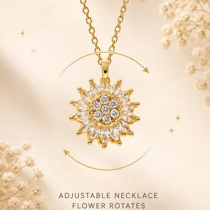 Op de afbeelding: Een goudkleurige ketting met een ronde hanger in de vorm van een bloem, bezet met heldere, marquise-geslepen stenen als bloemblaadjes en ronde stenen in het midden. De bloem kan draaien, aangegeven door een cirkelvormige pijl. De tekst luidt ADJUSTABLE NECKLACE FLOWER ROTATES.