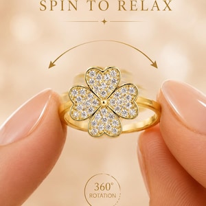 Könnte beinhalten: Ein goldfarbener Ring mit einem vierblättrigen Kleeblatt-Element, das mit klaren Steinen besetzt ist. Über dem Ring steht der Text SPIN TO RELAX, während ein gebogener Pfeil unter dem Kleeblatt eine 360-Grad-Drehung anzeigt.