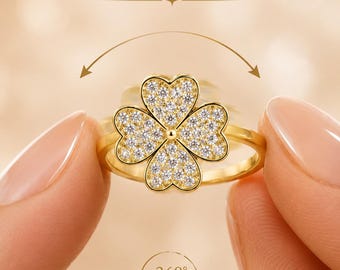 May include: A gold-toned ring featuring a four-leaf clover centerpiece encrusted with clear stones. The text SPIN TO RELAX appears above the ring, with a curved arrow indicating 360 degree rotation below the clover design.