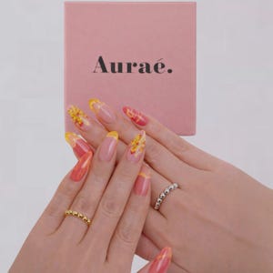 May include: A pink box with the Aurae brand name in black. Two hands showcase rings, one gold beaded and one silver beaded. The nails are long, with a French manicure design in yellow, orange, and pink.