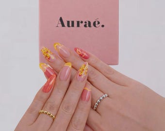 May include: Aurae pink box with the brand name in black. Two hands display rings, one gold beaded and one silver beaded. The nails are long, with a French manicure design in yellow, orange, and pink.