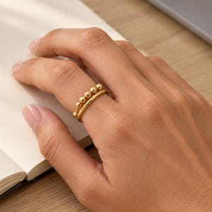 Peut inclure: Une bague dorée au design à double anneau. Un anneau est composé d'une série de petites perles sphériques lisses, tandis que le second anneau est un fil doré uni et poli. Les deux anneaux sont reliés sur les côtés.
