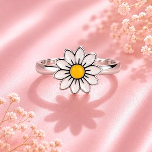 May include: A silver daisy ring. The ring features a daisy with white petals, a yellow centre, and black outlines. The ring is set against a pink background with floral accents, creating a delicate and feminine aesthetic.