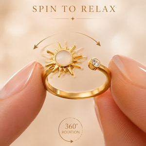 May include: A gold-toned ring featuring a sun-shaped charm with a central white stone and a separate small clear stone on the band. The text SPIN TO RELAX and 360 ROTATION are visible, along with a curved arrow indicating the sun charm rotates.