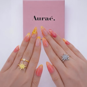 May include: Two hands display rings and manicured nails. One hand has a gold ring with a sun design and a diamond. The other hand has a silver ring with a sun design. The nails are painted with a gradient of yellow, orange, and pink.