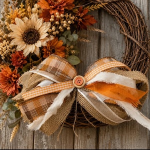 May include: A grapevine wreath decorated with artificial sunflowers, orange and rust-colored flowers, and greenery. A large bow made of burlap, plaid, and orange gingham ribbon is attached to the bottom, featuring a central wooden button detail.
