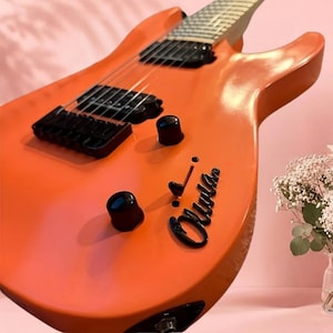 May include: An electric guitar with a bright orange body, black hardware, and black pickups. The body features a black script signature that reads Olivia. The guitar has a smooth finish, a black bridge, and two black control knobs on the front.