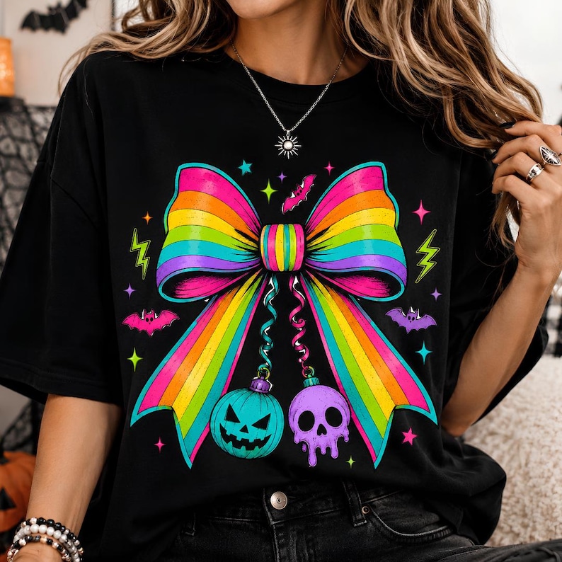 May include: A black t-shirt featuring a large, vibrant rainbow-colored bow graphic. Hanging from the bow are a teal jack-o'-lantern and a purple skull. The design is accented with small pink and teal bats, stars, and lightning bolt shapes.