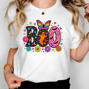 May include: A white t-shirt featuring a colorful graphic print of the word BOO. The design includes a rainbow butterfly, an orange ghost, pink checkered patterns, bats, candies, and various bright flowers, stars, and hearts surrounding the letters.