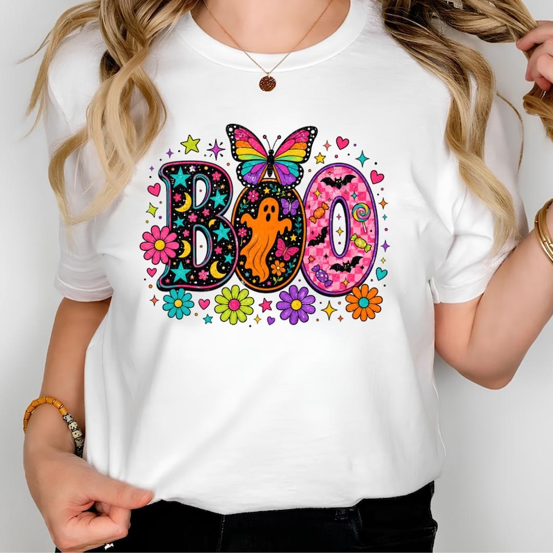May include: A white t-shirt featuring a colorful graphic print of the word BOO. The design includes a rainbow butterfly, an orange ghost, pink checkered patterns, bats, candies, and various bright flowers, stars, and hearts surrounding the letters.