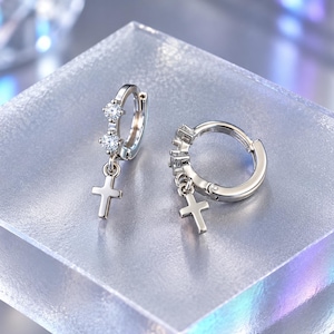 May include: A pair of silver-toned hoop earrings featuring two small, clear, round-cut stones set along the front of each hoop, with a small, plain silver-toned cross charm dangling from the bottom of each earring, creating a cohesive set.