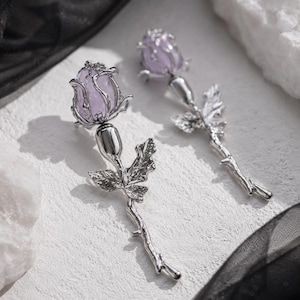 May include: A pair of silver-toned earrings shaped like long-stemmed roses. Each earring features a light purple, translucent, rounded stone set within a metallic rosebud frame, attached to a textured silver stem with detailed leaves and thorns.