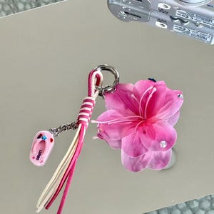May include: A keychain featuring a large, translucent pink flower with small embedded crystals, a pink and white striped leather tassel, and a small pink flip-flop charm with the text LURSD printed on it, all attached to a silver metal ring.