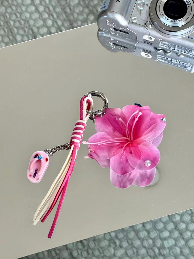 May include: A keychain featuring a large, translucent pink flower with small embedded crystals, a pink and white striped leather tassel, and a small pink flip-flop charm with the text LURSD printed on it, all attached to a silver metal ring.