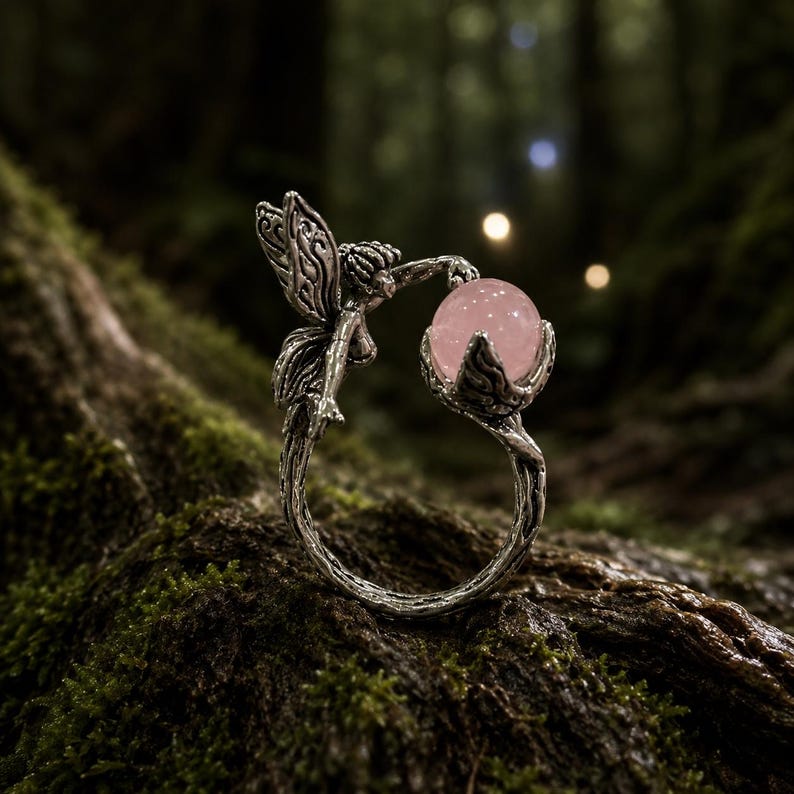 May include: A silver-toned ring featuring a textured band resembling a tree branch, topped with a small winged figure reaching towards a smooth, round, pale pink gemstone held within a leaf-shaped setting at the opposite end of the open band.