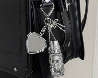 May include: A silver-toned metal keychain accessory featuring a heart-shaped compact mirror and a small glass perfume bottle encased in a decorative silver chain mesh. The charm set includes a small bow detail and attaches to a black bag.