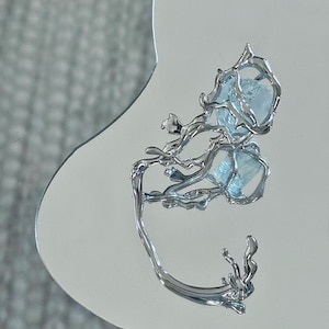 May include: A silver-toned metal ear cuff featuring an organic, branch-like structure that wraps around the ear. The design incorporates a single, pale blue, translucent, raw-cut gemstone held within the center of the metallic framework.