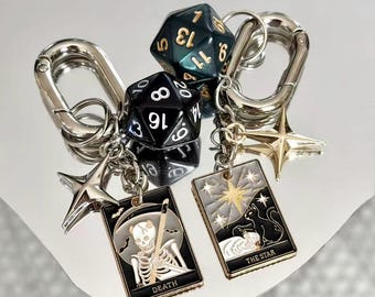 May include: Two silver-toned metal keychains featuring polyhedral dice, star charms, and rectangular enamel pendants. One pendant depicts a skeleton with the text DEATH, while the other shows a cat under a star with the text THE STAR.