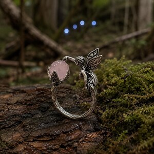 May include: A silver-toned metal ring featuring a textured band that resembles a tree branch. One end of the band depicts a winged figure, while the other end holds a smooth, round, pale pink gemstone within a small, petal-like setting.